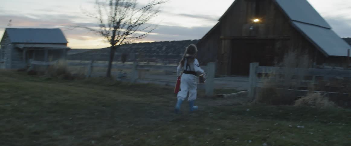 A young girl in boots and coveralls runs through a gate towards a barn at sunset
