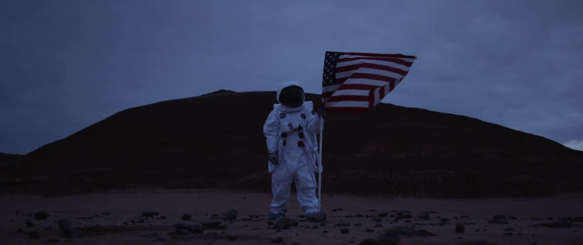 A person wearing a space suit stands in front of a hill holding a flag waving on a small pole