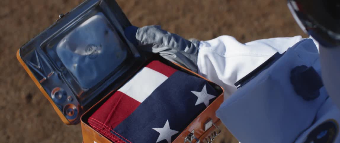 A person in an astronaut suit opens a case with a flag in it
