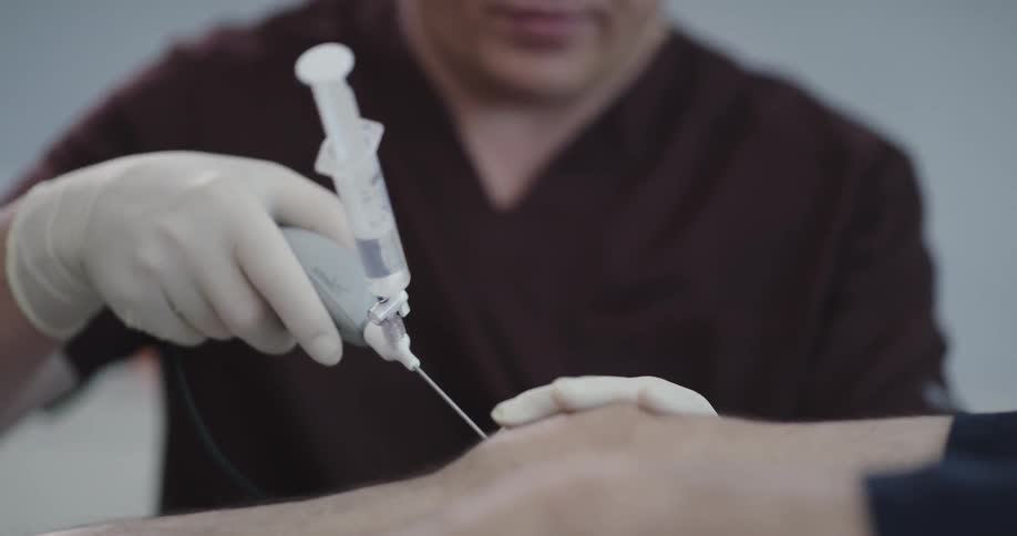 A medical professional guides a large needle into the knee of a patient who is reclined in front of him