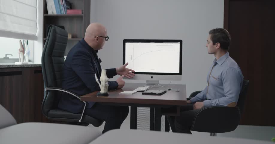 Two men siton opposite sides of a desk while the one man shows the other something on a computer screen