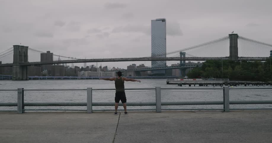 A man running and stretching along a river walkway across from a large city