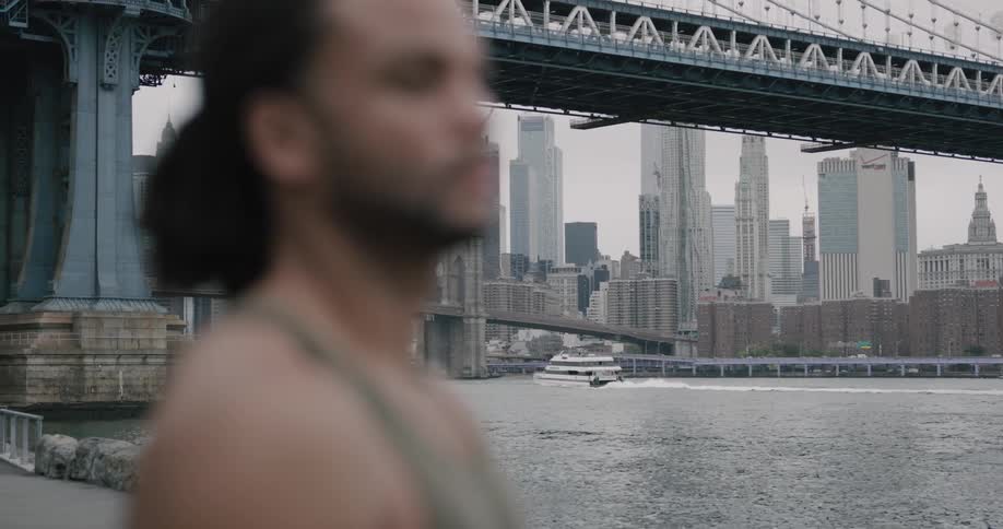 A man stands in front of a suspension bridge near some water