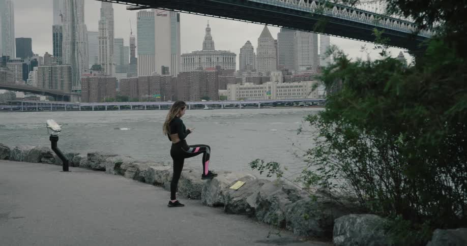 Woman holding smartphone runs along on the promenade and takes a break to wear bluetooth earphones next to the river across the city on a cloudy day