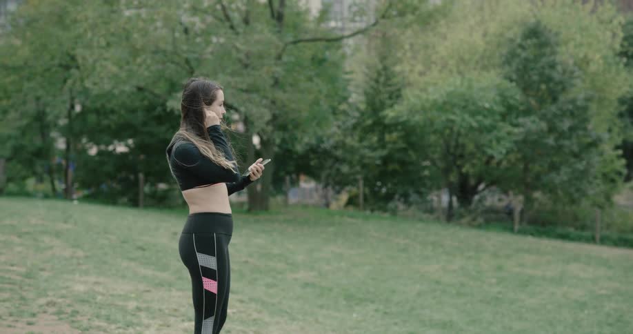Woman holding smart phone stands on the grass and wears earphones during daylight