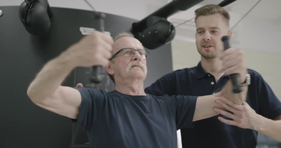 An older man is performing a chest exercise with a pulley based piece of equipment under the supervision of a physical therapist