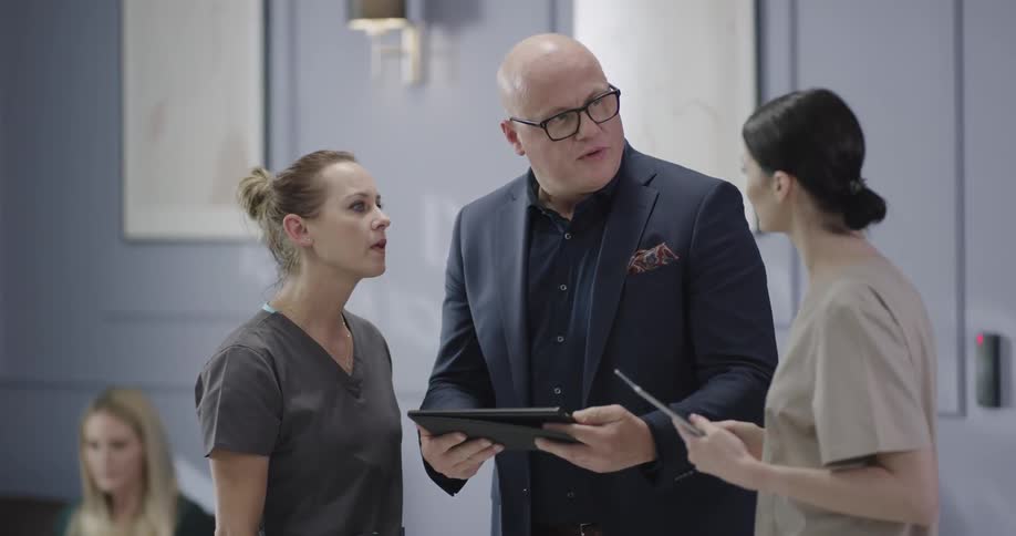 A bald man in a suit coat is looking at a smart tablet and discussing it with a female associate when he is approached by another woman in medical scrubs