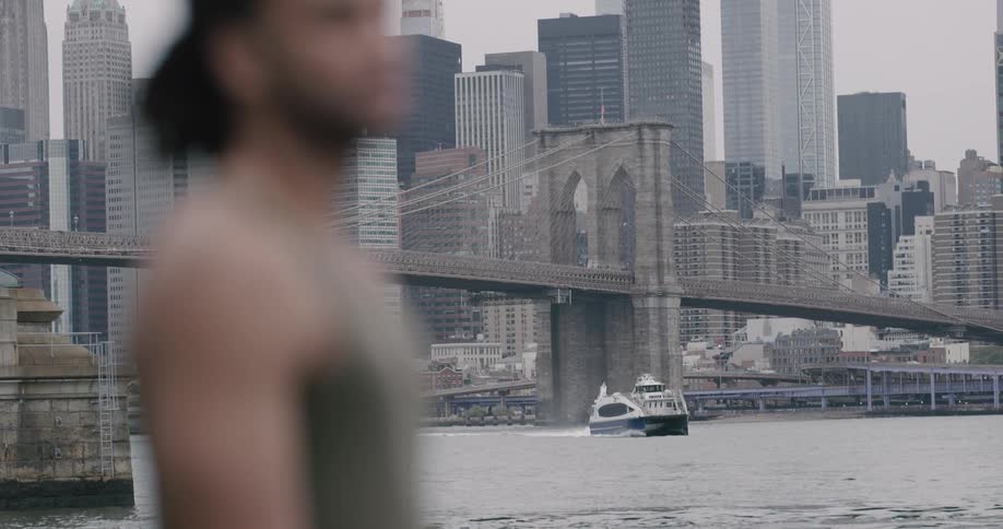 A man stands on a shore near the water looking out at a bridge over the water with a boat going underneath and a huge city with many sky scrapers