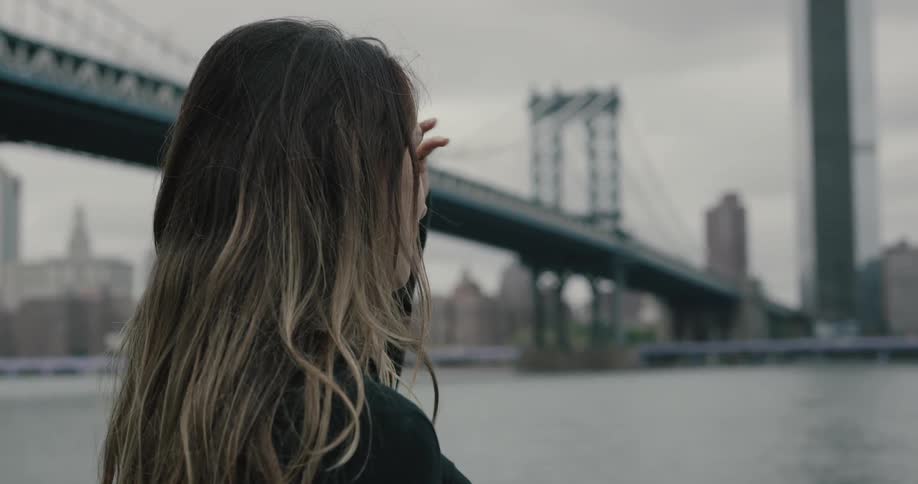 A woman fluffs her hair with her hand while holding a cell phone and looking out at the city harbor and bridge