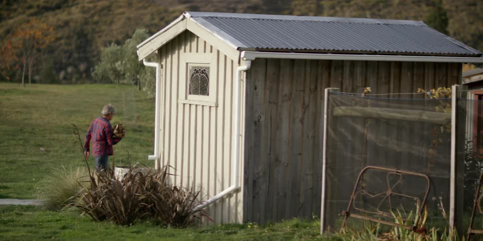 A man carries a pile of wood past a shed in a country setting