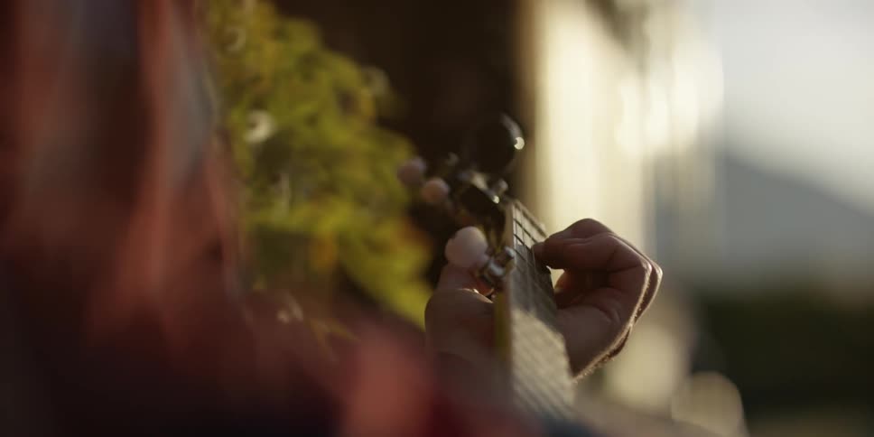 A man sitting on a bench next to a house plays a guitar