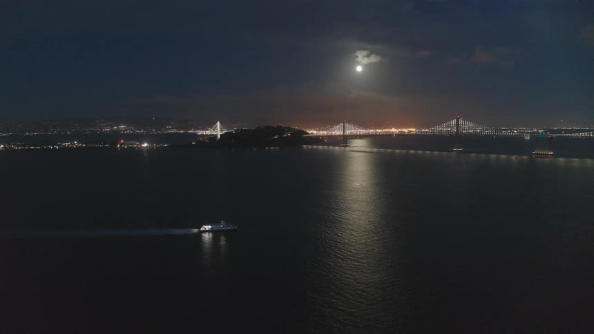 The moon glows in the night sky above a bridge and a boat