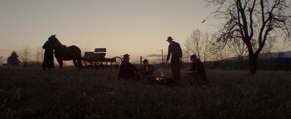 Several men sit around a camp fire as the sun sets while another tends to a horse