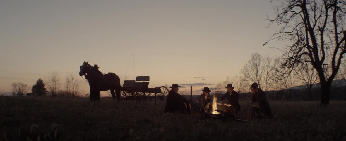 Four people sit around a campfire while someone tends to a horse nearby