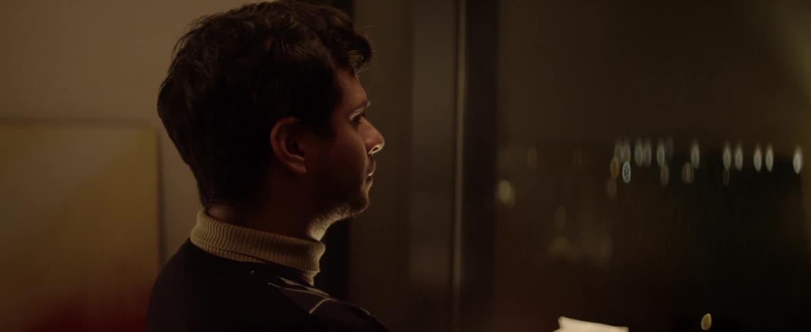 A man in a white turtleneck is standing near a wall