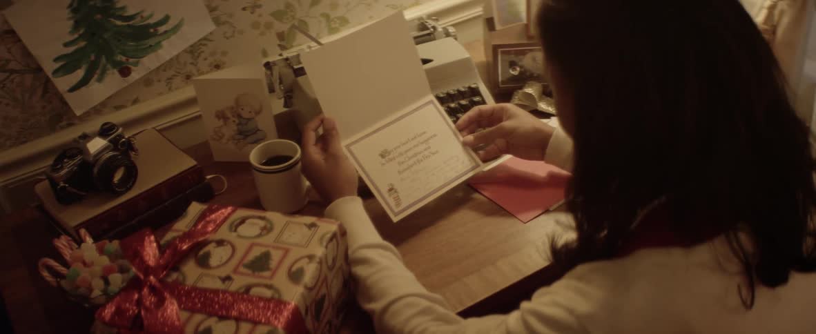 A woman sits down at a desk with a typewriter and christmas gift to look at a christmas card