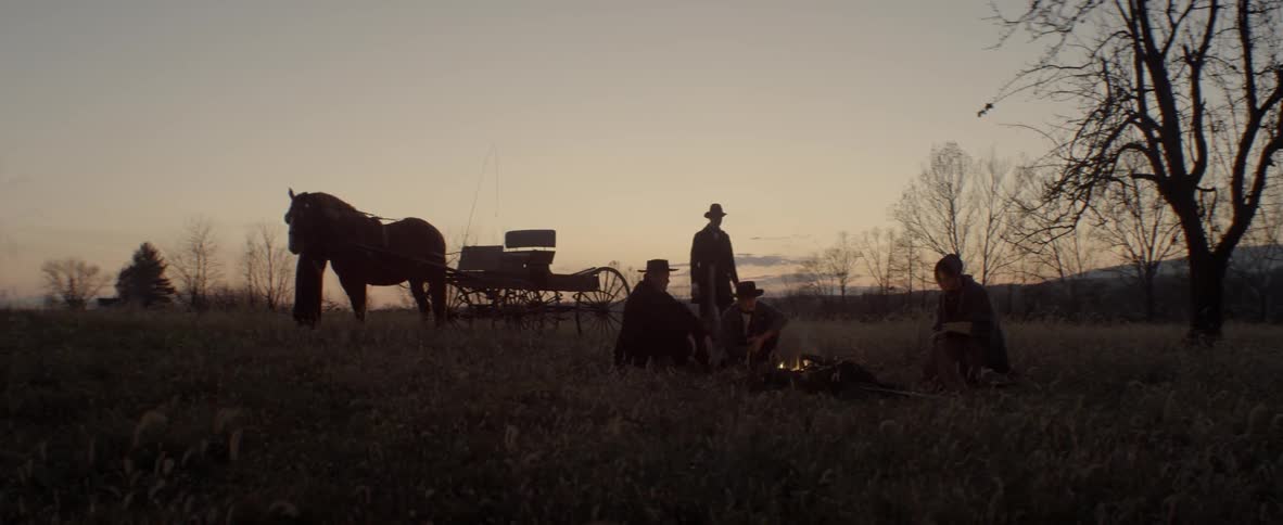 Three people sitting around a campfire as a fourth loads a hoe onto a horse drawn wagon