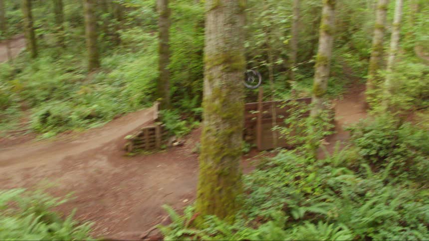 A male wearing a helmet rides a mountain bike up a ramp and lands a large jump as he rides through a bike course in the forest