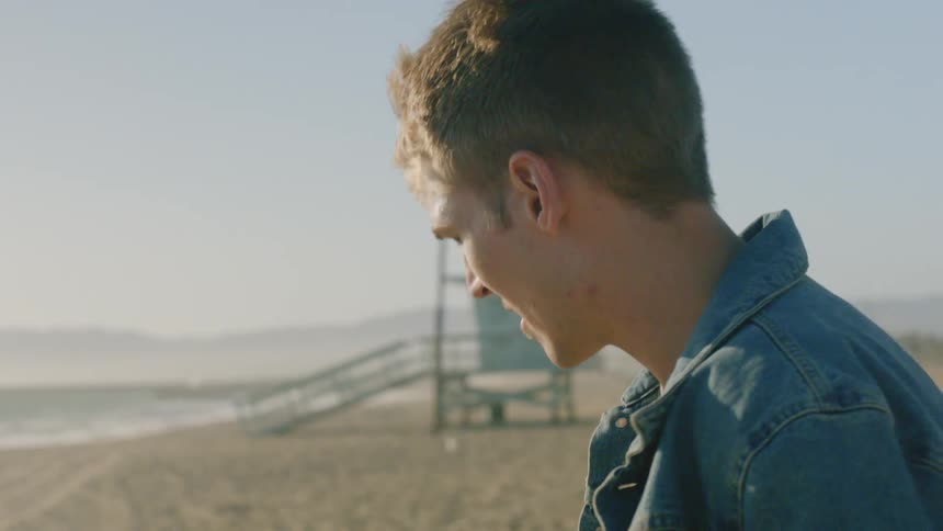 Blonde man wearing denim jacket walks on the white sand beach on a sunny day next to the ocean