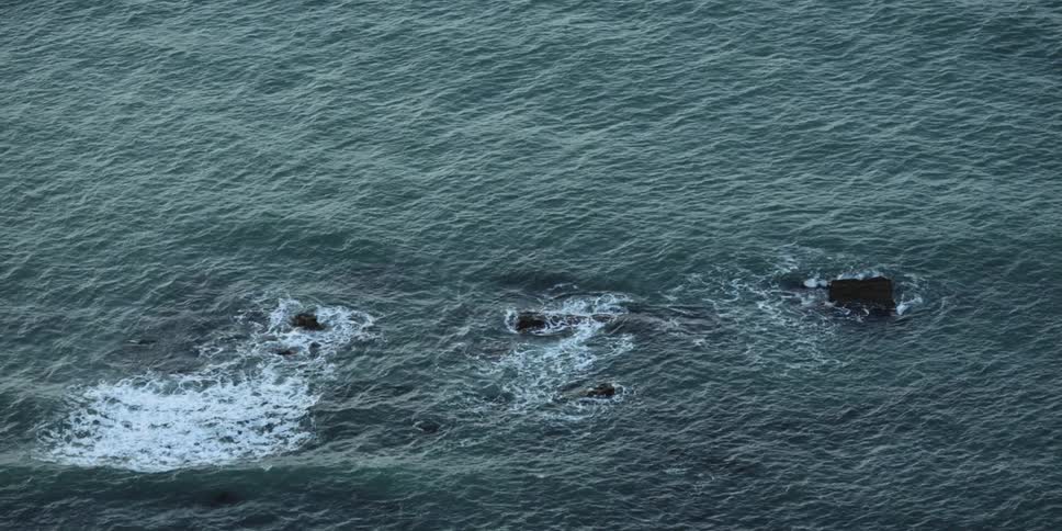 The tips of a small cluster of jagged rocks protrudes from the ocean surface as small waves wash around and over them