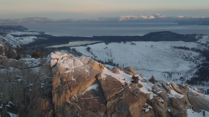 A rocky cliff with snow with a field and a lake in the background