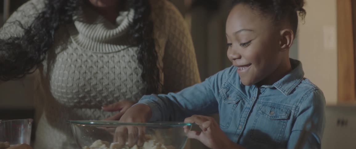 A young black girl is mixing ingredients in a glass bowl using her hand while her mother hands her an egg