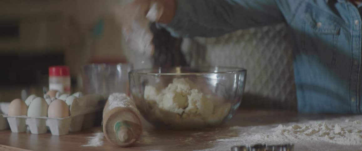 A person spreads flour on a rolling pin then grabs a handful of dough from a mixing bowl while another person watches from behind