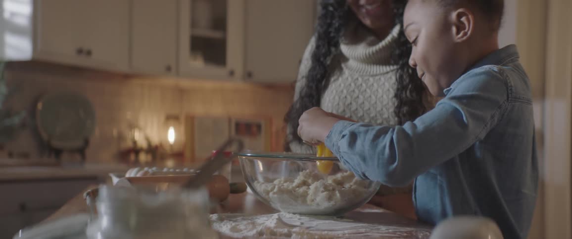 A woman watching a girl break an egg into a glass bowl of dough in a kitchen