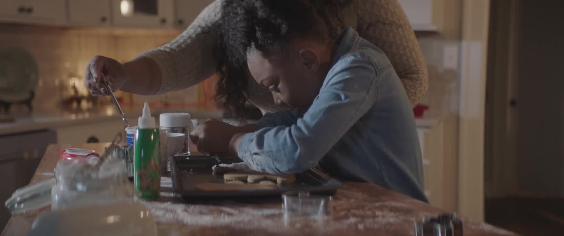 A young girl is decorating gingerbread man cookies with her mother in the kitchen