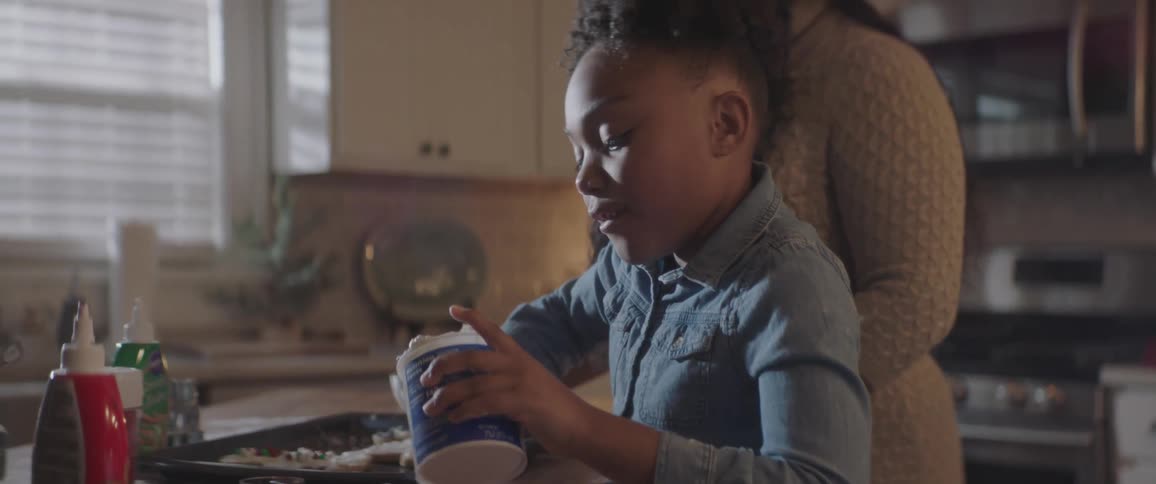 A girl scooping icing and spreading it on cookies as a woman watches over her
