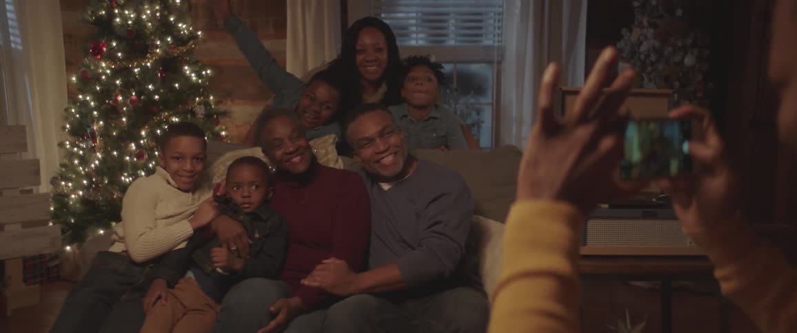 Black man takes picture of black family sitting on the couch next to the christmas tree with a smartphone at home