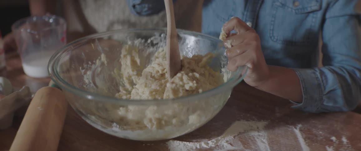 A child isusinga wooden spoon to stir cookie dough in a large glass bowl on a counter in a kitchen