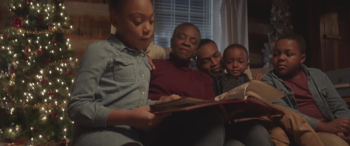 Grandparents sit on a couch with their three grandchildren reading a book at christmas