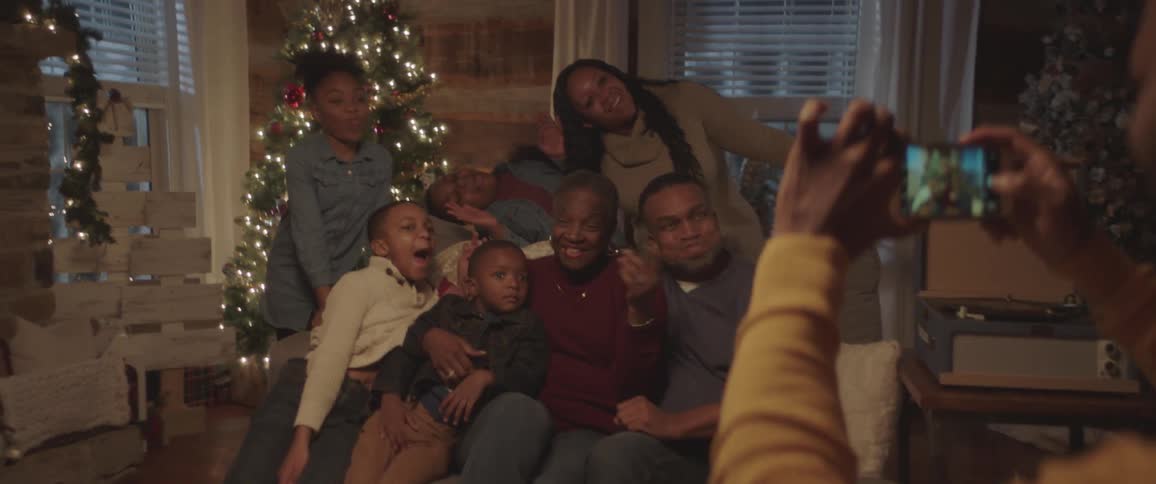 Black man takes picture of black family sitting on the couch next to the christmas tree with a smartphone at home