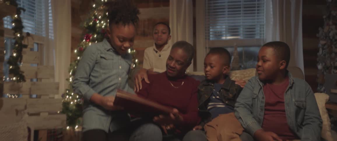 A young girl walks towards a couch where her family sits to show them a book