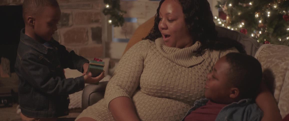 A young boy brings a christmas gift box to his mother sitting on the couch