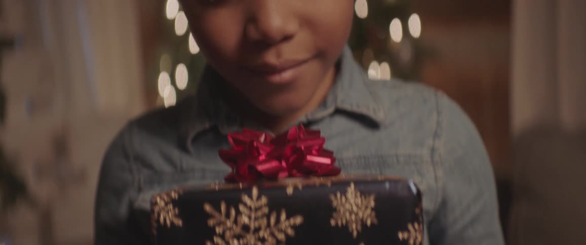A little girl holding a gift wrapping in black paper with golden snowflakes smiles into the camera