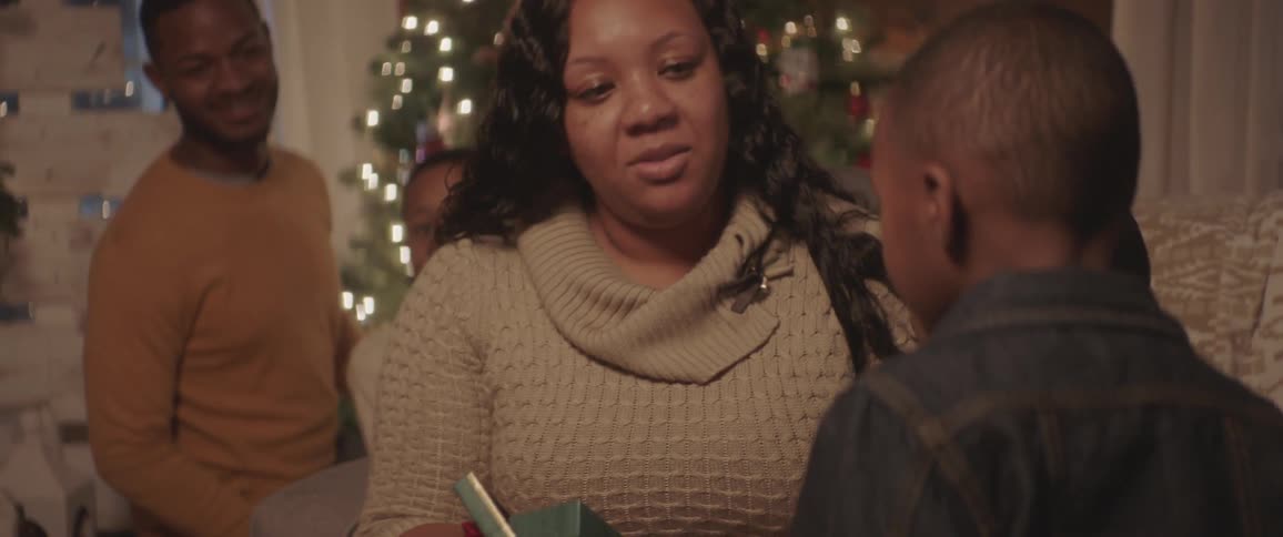 A woman opens a present in front of a christmas tree and then hugs one of her two young sons who are sitting next to her