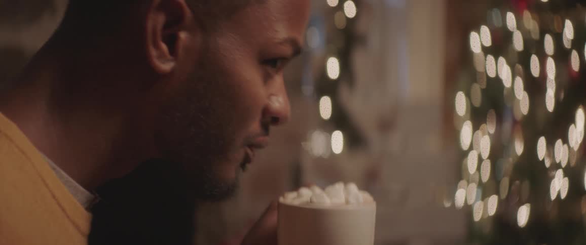 A man drinks hot chocolate with marshmallows out of a mug in front of a christmas tree