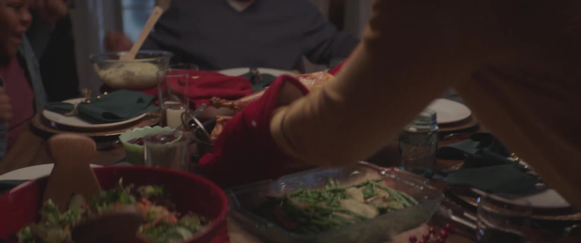 A black man sets a roasted turkey on a table with all the trimmings while the rest of the family cheers