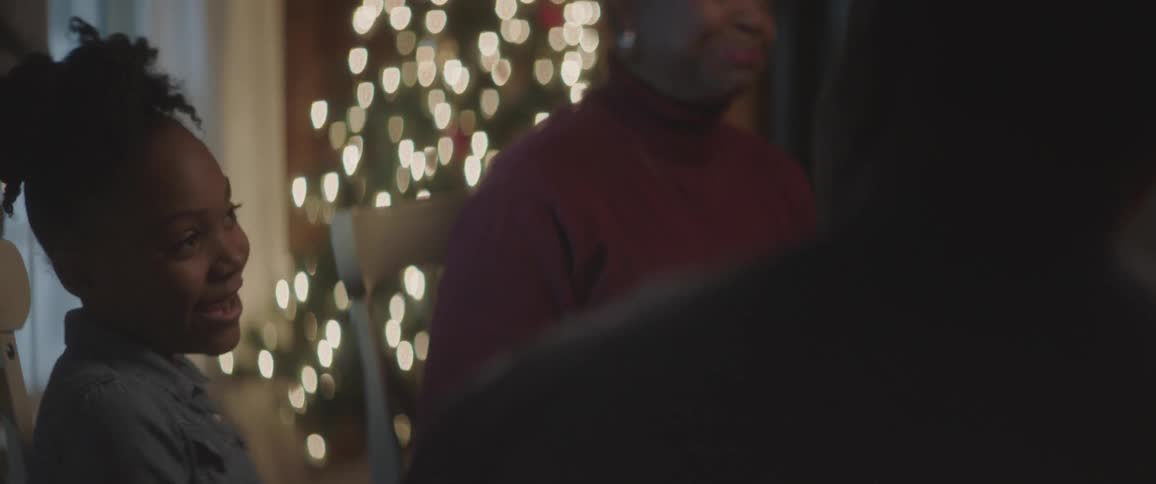 A young black girl is talking to a man and older woman at a table with a christmas tree lit up in the background