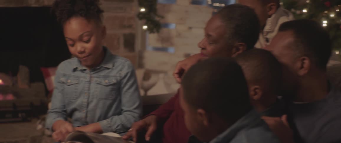 A young girl reads a book out loud while her father grandfather and siblings sit nearby