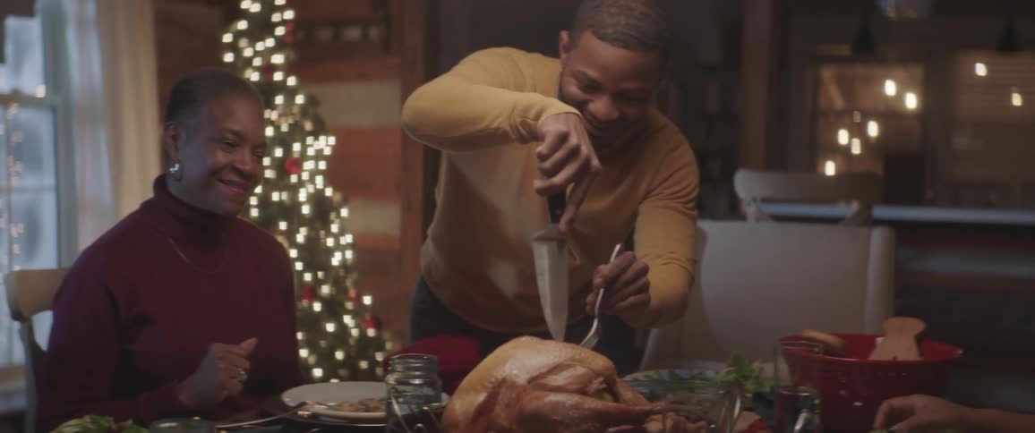 A man tries to cut a large bird for a meal but passes the knife to another person