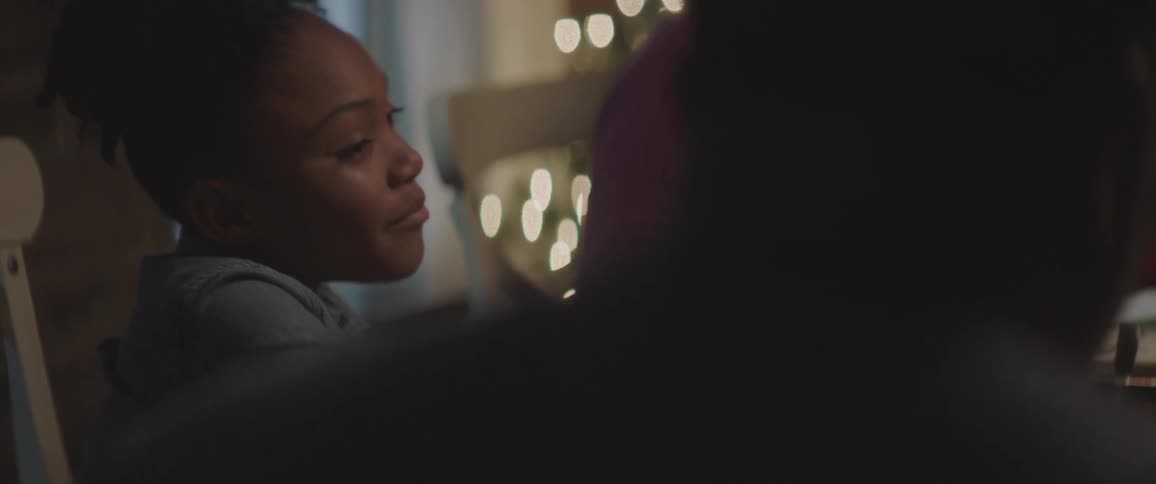 A girl sits at the table and talks to her family with a lit christmas tree in the background