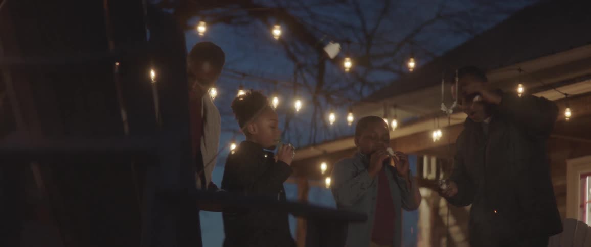 Two adults and two children are roasting marshmallows and eating smores near a fire and beneath string lights