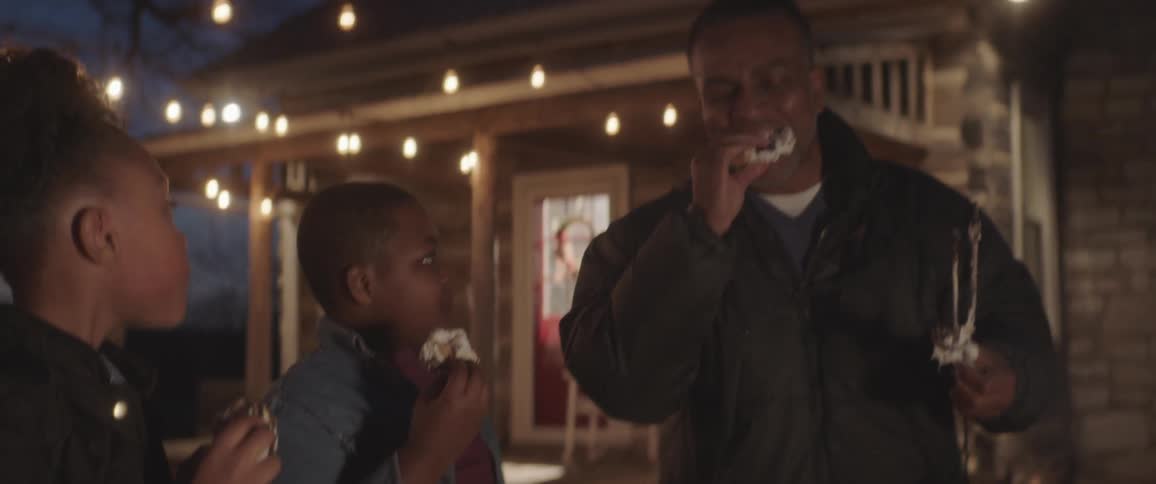 A man and two children toast marshmallows on a fire outside of a quaint house