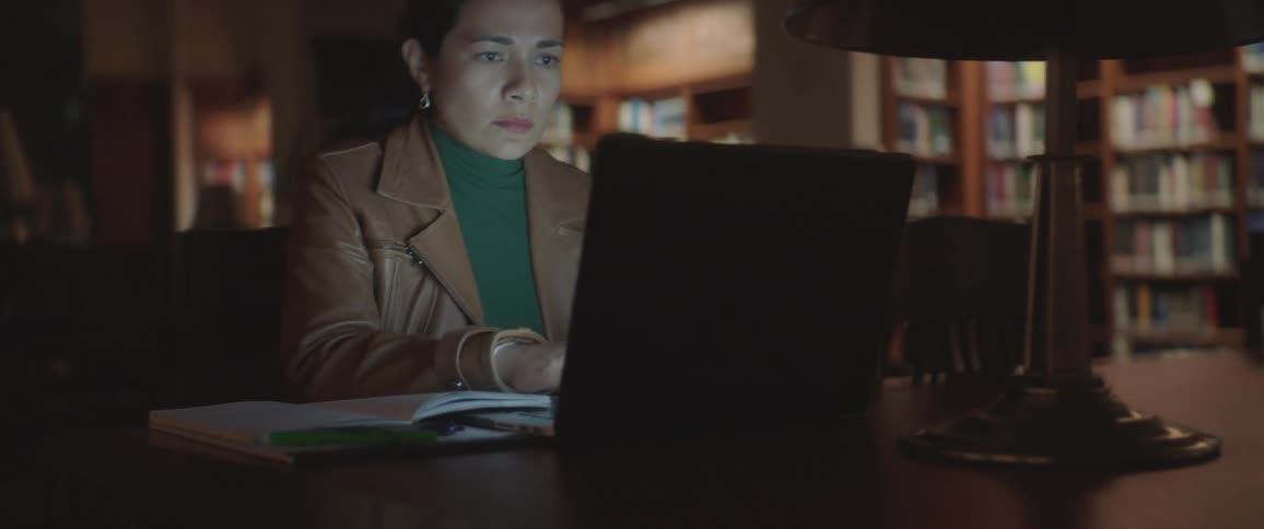 Woman sitting at the table uses laptop in the library at night