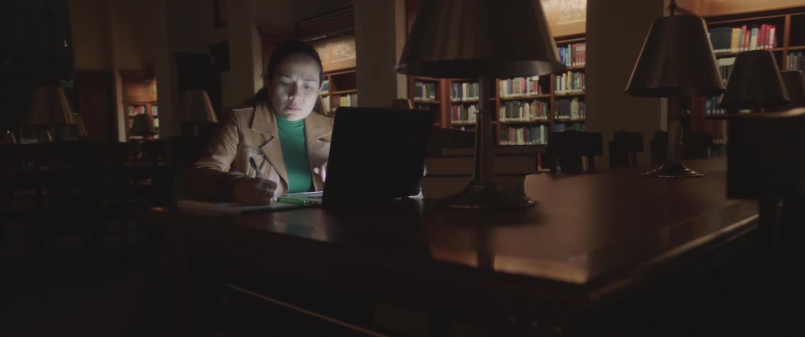 Woman sits at table in dark library taking notes
