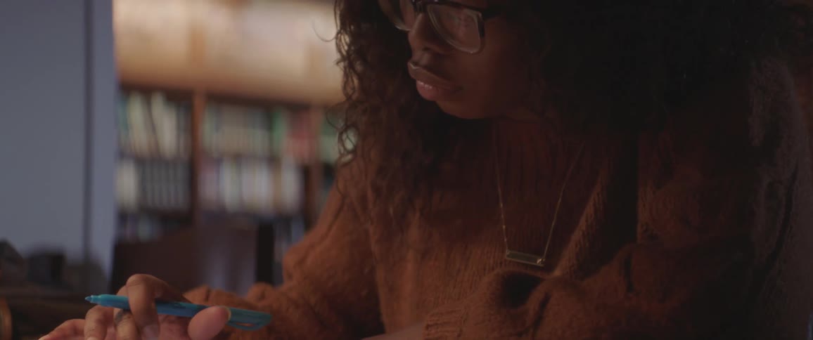 A young woman with glasses and a brown sweater does her homework in a library