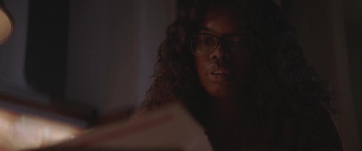 A young black woman has a look of frustration on her face as she looks over pages in a textbook in a darkened room